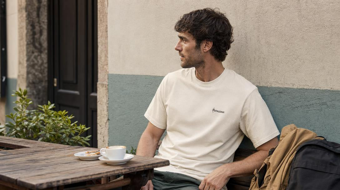Un uomo indossa una T-shirt Patagonia mentre si siede a un tavolino all’aperto, con una giacca e uno zaino accanto per una giornata in città.