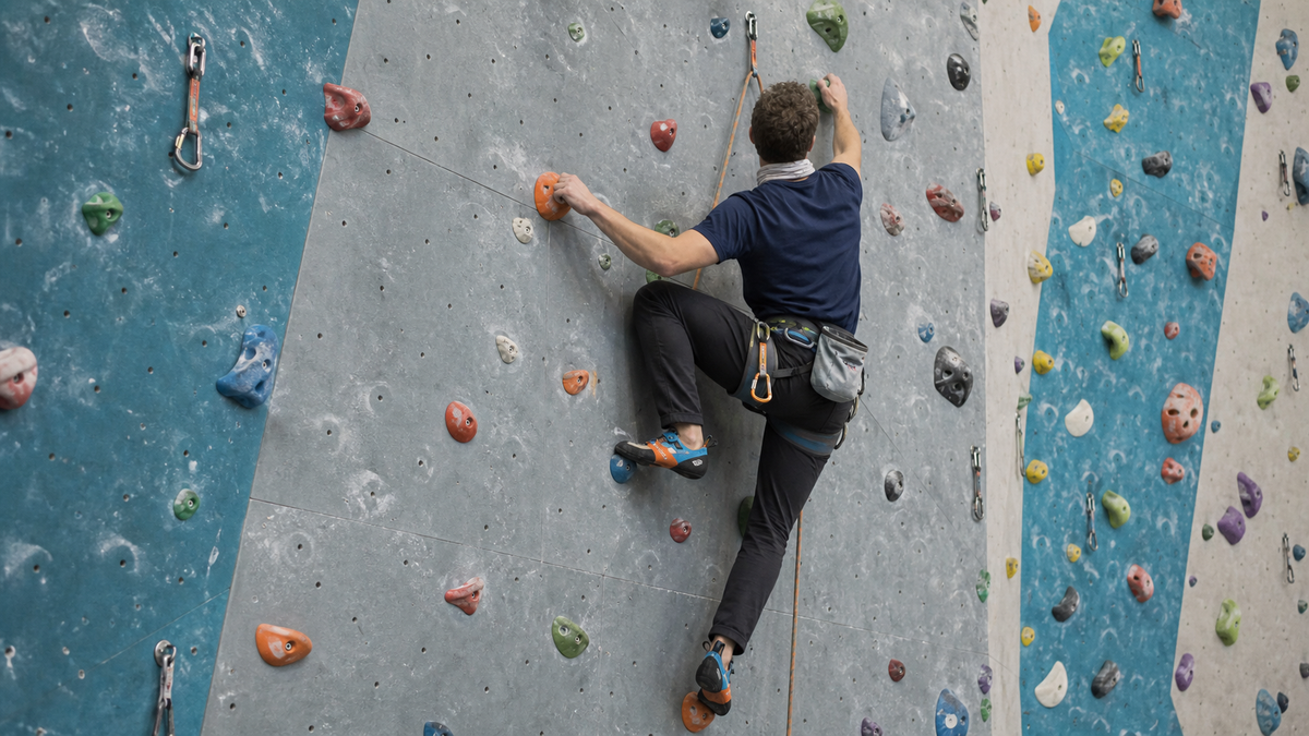 Arrampicatore su parete indoor con prese, maglia tecnica blu, scaldacollo bianco, scarpette arancioni e moschettone
